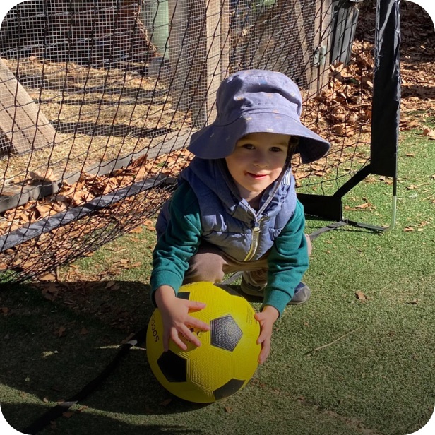 A child in a sun hat crouching to pick up a yellow ball