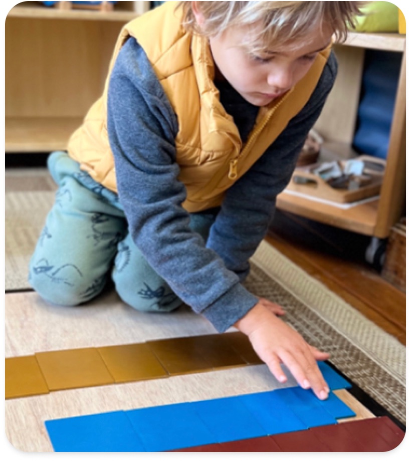 A boy in a yellow vest unrolling a work mat on the classroom floor